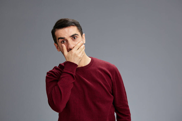 hombre guapo cubre la boca con gesto de la mano posando con las manos suéter rojo aislado fondo - Foto, Imagen