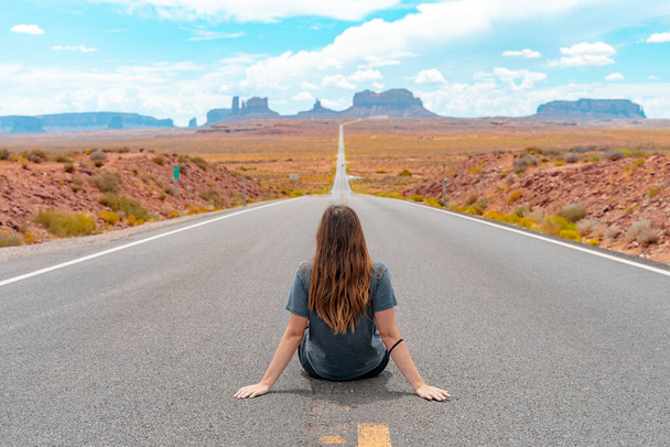 Attractive young woman from back view sitting in the middle of American straight road in the desert. - Foto, Imagem