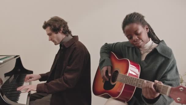 Lento medio de talentosa pareja interracial de músicos tocando piano y guitarra juntos en interiores - Metraje, vídeo