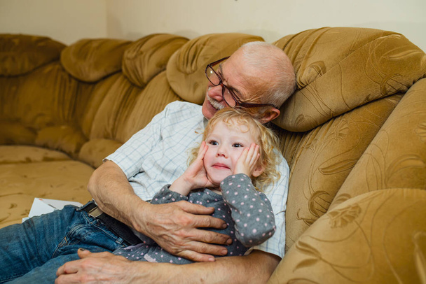 祖父は孫がソファに座っているのを抱きます。家にいる老人は女の子と遊ぶ。隔離後の老人と少女の出会い - 写真・画像