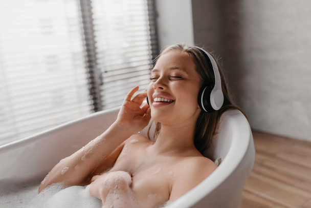 Senhora relaxada descansando em banho quente com os olhos fechados e ouvindo música em fones de ouvido sem fio, espaço de cópia - Foto, Imagem