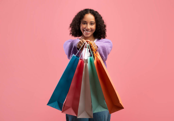 Szép fiatal fekete nő gazdaság shopper táskák kinyújtott kézzel rózsaszín stúdió háttér - Fotó, kép