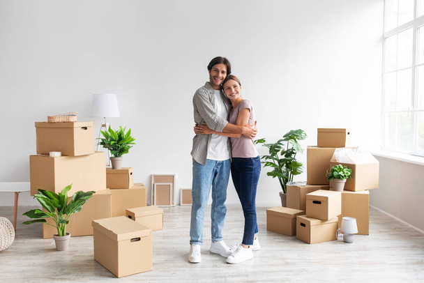 Feliz joven caucásico hombre y mujer en abrazos casuales en la habitación con cajas de cartón, alquiler de piso y reubicación - Foto, imagen