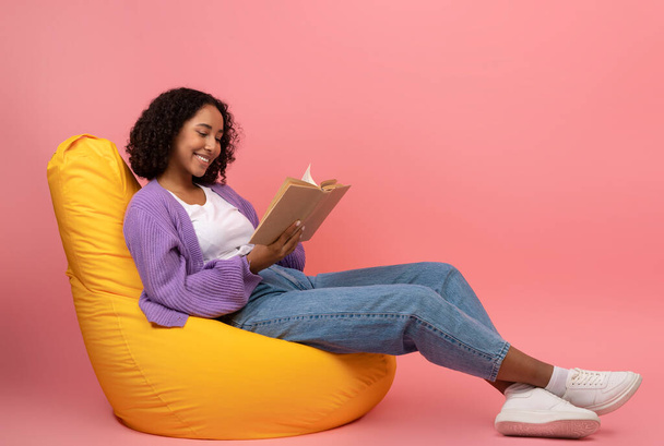 Relajada joven mujer negra sentada en la silla de la bolsa de frijol con libro abierto sobre fondo de estudio rosa, espacio en blanco - Foto, Imagen