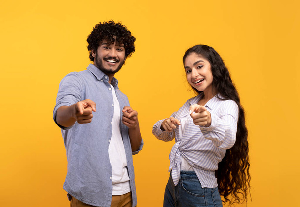 Και τι γίνεται με σένα. Ενθουσιασμένο ζευγάρι Ινδιάνων να σημαδεύει με δείκτες την κάμερα, ποζάροντας πάνω από κίτρινο φόντο στούντιο - Φωτογραφία, εικόνα