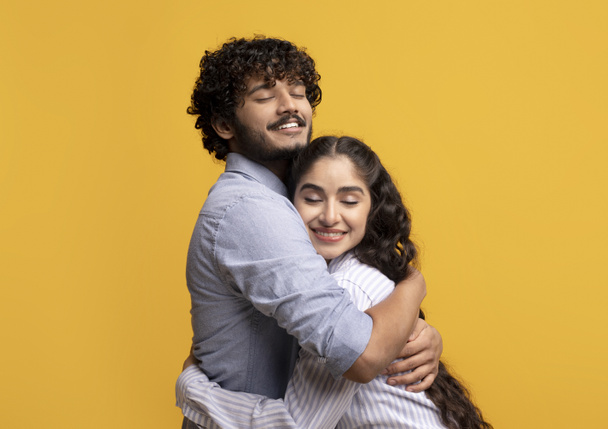 Portrait of affectionate indian couple hugging with closed eyes while standing isolated over yellow studio background - Photo, Image