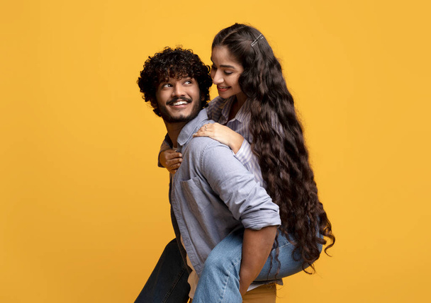 Pretty indian lady piggybacking her boyfriend, smiling guy carrying his girlfriend on his back, yellow background - Photo, Image
