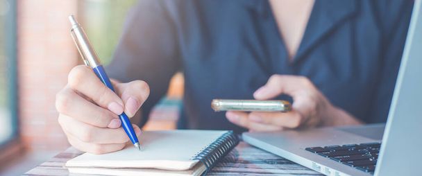 Woman hand writing on a notepad with a pen and use a smartphone in the office.for web banner. - Foto, imagen