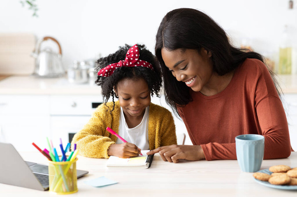 Портрет заботливой чёрной мамы, помогающей своей маленькой дочери с домашней работой - Фото, изображение