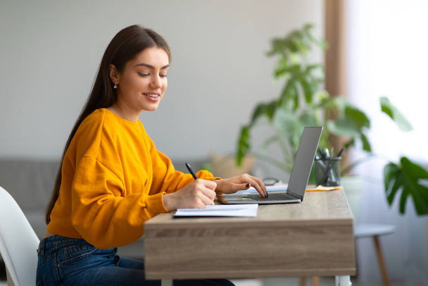 Happy Caucasian woman using laptop pc, working or studying online, taking notes during webinar or remote lesson at home - Photo, Image