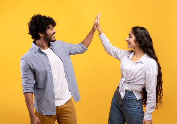 Common success. Young excited indian couple giving high-five, clapping hands, greeting each other over yellow background - Photo, Image