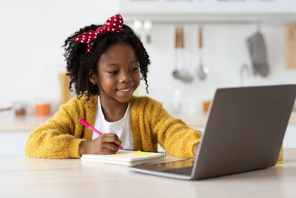 Linda chica negra preadolescente escribiendo en bloc de notas mientras estudia en línea con el ordenador portátil - Foto, imagen