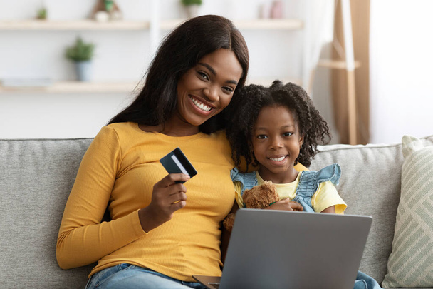 Paiements en ligne. Mère noire et enfant femelle avec ordinateur portable et carte de crédit - Photo, image