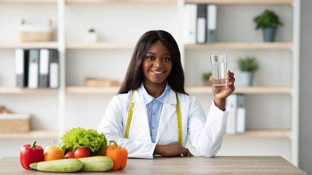 Mladá žena africký americký nutriční pracovník drží sklenici vody, zatímco sedí u stolu s ovocem a zeleninou - Fotografie, Obrázek