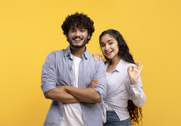 Portrait eines glücklichen indischen Paares, das sich umarmt, Frau zeigt okay Geste und lächelt in die Kamera über gelbem Hintergrund - Foto, Bild