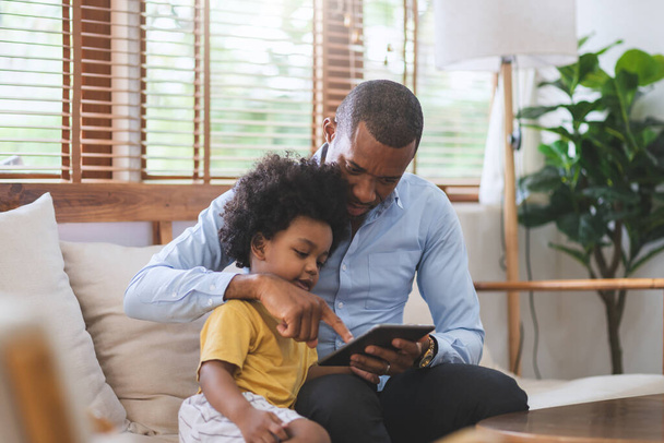 Afro-Amerikalı baba ve küçük oğlu evdeki oturma odasında dijital tablet kullanıyorlardı. İnternet teknolojisiyle mutlu siyah aile. - Fotoğraf, Görsel