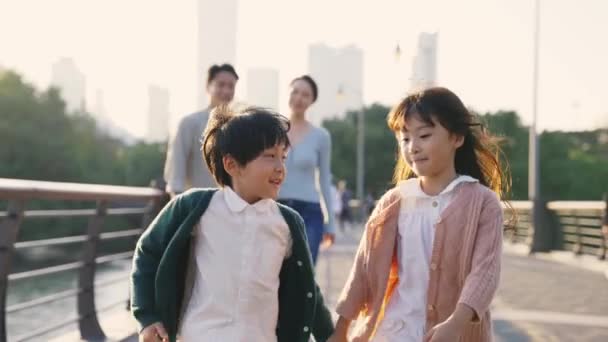 kaksi onnellista aasialaista lasta kävelemässä puistossa vanhempien kanssa taustalla - Materiaali, video