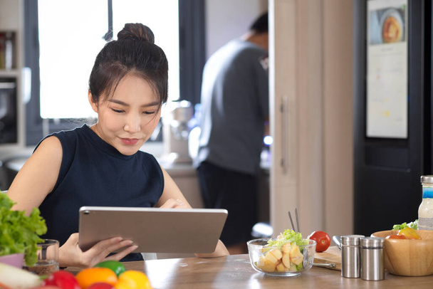 Aziatisch jong vrouw met behulp van draagbare tablet aan haar huis eettafel, terwijl haar vader het bereiden van voedsel in de keuken. - Foto, afbeelding