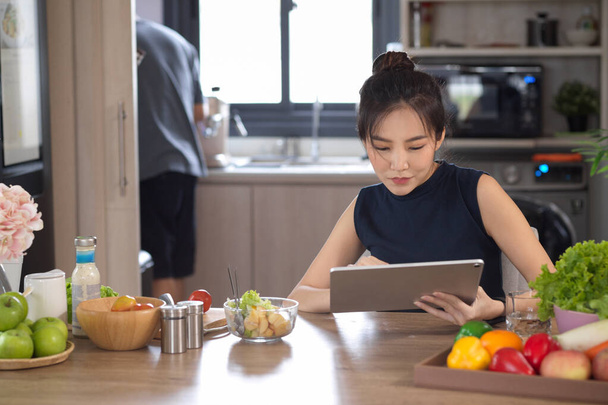 A cheerful Asian young woman sits at her dining table and uses her portable tablet touchpad while eating salad for breakfast. - Photo, Image