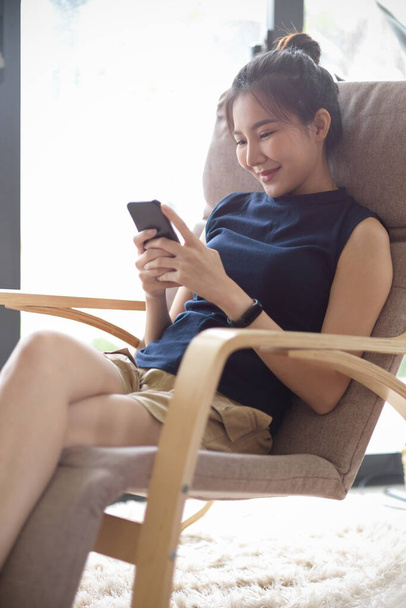 Ritratto, attraente asiatica teen donna si siede in poltrona moderna e confortevole in luminoso soggiorno, utilizzando smartphone, godere di chiacchierare con i suoi amici. - Foto, immagini