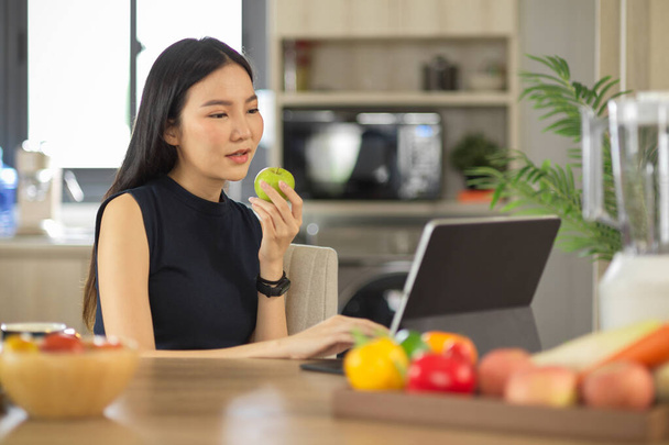 Aantrekkelijke Aziatische vrouw die groene appel eet terwijl ze iets online leest via een draagbare tablet in de keuken. Gezond levensstijl concept - Foto, afbeelding