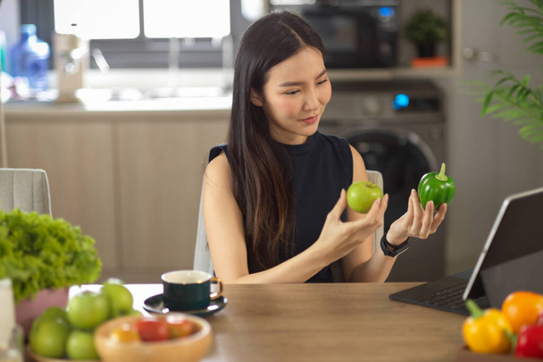 Charmante femme asiatique regardant l'écran de la tablette et tenant une pomme verte et un poivre vert dans la cuisine, Apprentissage de la cuisine en ligne, blogueur cuisine. - Photo, image