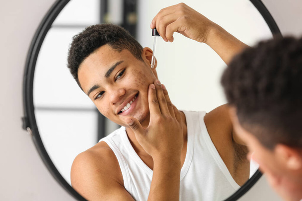 Молодой афроамериканец использует сыворотку для ухода за кожей в ванной комнате - Фото, изображение