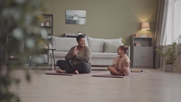 Amplio slow mo de niña afroamericana de 5 años y su madre sentadas en colchonetas de yoga en salón meditando juntas - Imágenes, Vídeo