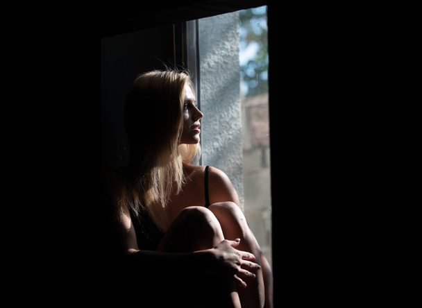 Sensual girl near window. Woman in shadow. Female model with shadows on face looking seductive and sensual on dramatic black studio with light. - Photo, image