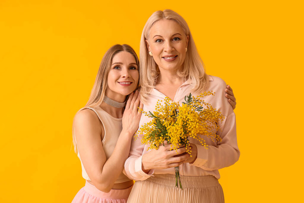 Νεαρή γυναίκα με τη μητέρα της και λουλούδια μιμόζα σε κίτρινο φόντο - Φωτογραφία, εικόνα
