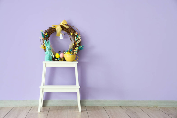 Όμορφο στεφάνι Πάσχα, κουνέλι και κύπελλο για σκαμνί στέπα κοντά στο χρώμα τοίχο στο δωμάτιο - Φωτογραφία, εικόνα
