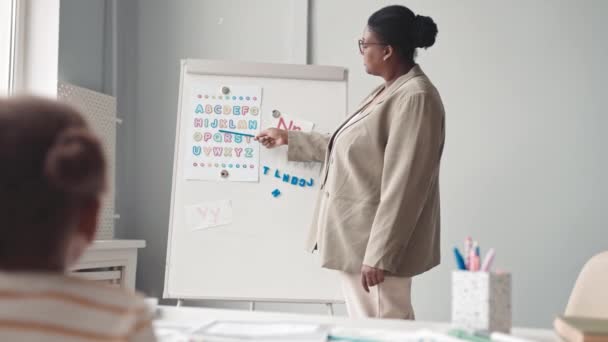 Ralentissement moyen de l'enseignante afro-américaine debout au tableau blanc pointant vers les lettres de l'alphabet anglais tout en donnant des leçons à une fille intelligente préscolaire assise au bureau devant elle - Séquence, vidéo