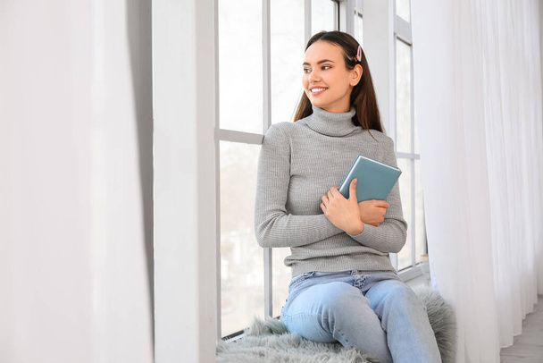 Pretty young woman with book sitting on window sill - 写真・画像
