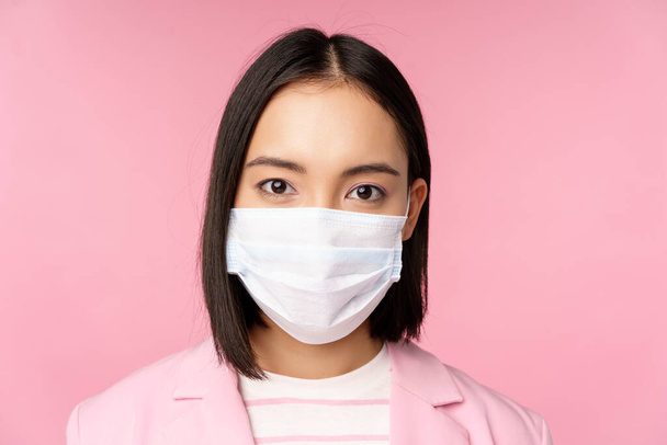 Primer plano retrato de mujer de negocios japonesa en la máscara de la cara médica, traje, mirando a la cámara, de pie sobre el fondo rosa - Foto, Imagen