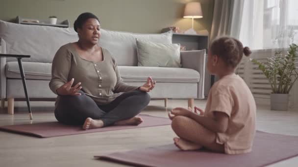 Lento de cuerpo joven mujer afroamericana positiva enseñando a su hija de 5 años de edad meditación sentada en pose de loto en colchonetas de yoga en casa - Imágenes, Vídeo