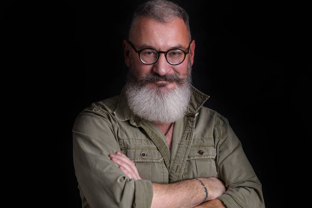 Mature bearded caucasian man looks slyly at camera with eyeglasses on his face, military style green shirt, black background - Fotó, kép