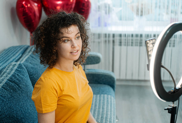 Curly haired woman recording video on mobile phone with balloons hearts at home - Fotó, kép