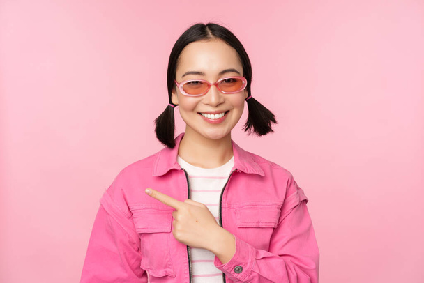 Retrato de chica asiática sonriente en traje elegante, gafas de sol, señalando el dedo a la izquierda, mostrando publicidad, pancarta, de pie sobre fondo rosa - Foto, Imagen