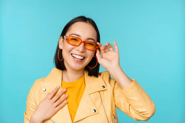 Close up retrato de mulher asiática elegante em óculos de sol, rindo e sorrindo, olhando feliz, posando em roupas da moda sobre fundo azul - Foto, Imagem