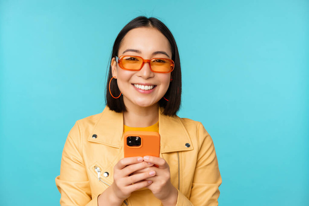 Portrait of enthusiastic asian woman in sunglasses, using mobile phone, smiling and laughing, looking happy, holding smartphone, standing over blue background - Fotó, kép