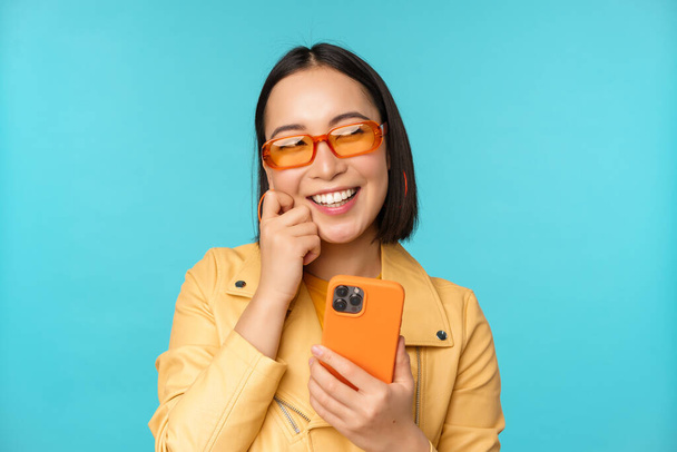 Счастливая стильная девушка, пользующаяся смартфоном и смеющаяся, улыбающаяся камере, стоящая на голубом фоне - Фото, изображение