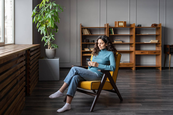 Молодая женщина сидит в уютном офисе или дома сидит в кресле просматривая социальные сети на смартфоне - Фото, изображение