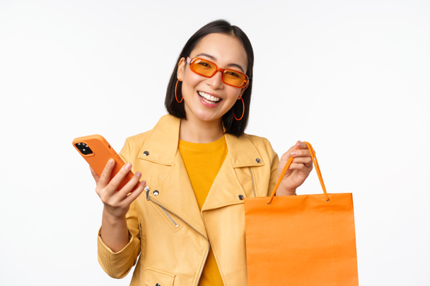 Stijlvolle Aziatische meisje shopper, draagt een zonnebril, het houden van boodschappentas en smartphone, gaan voor kortingen in de winkels, staande over witte achtergrond - Foto, afbeelding
