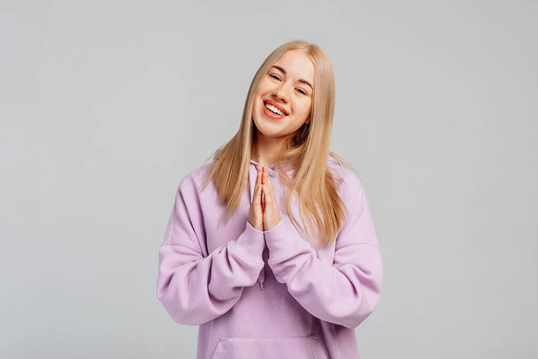 Sonriente mujer joven de pelo rubio con capucha violeta se levanta sobre fondo gris se toman de la mano en la oración Namaste gesto, mira a la cámara se sienten agradecidos. Concepto de lenguaje corporal - Foto, imagen