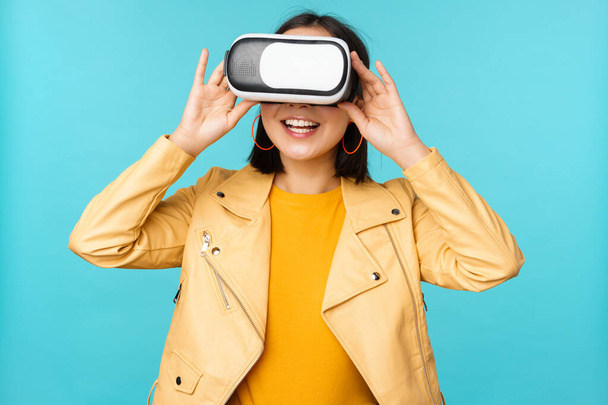 Портрет счастливой азиатской модели с помощью гарнитуры VR, улыбающейся и смеющейся в очках виртуальной реальности, стоящей на синем фоне - Фото, изображение