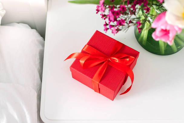 Kimppu tulppaanit ja punainen lahjapaketti yöpöydällä lähellä sängyn makuuhuoneessa. Käsite naisten päivä, äitien päivä - Valokuva, kuva