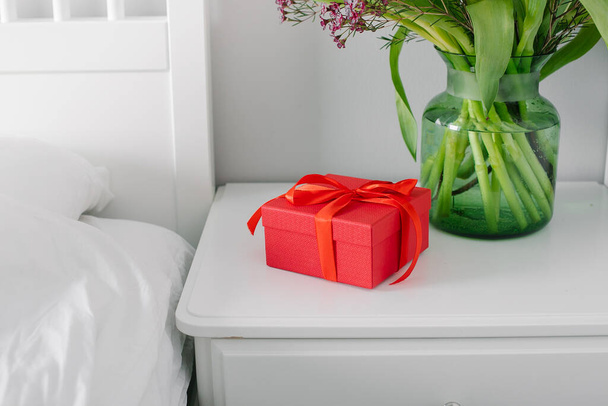 寝室のベッドの近くのナイトスタンドのチューリップと赤いギフトボックスの花束。女性の日、母の日の概念 - 写真・画像