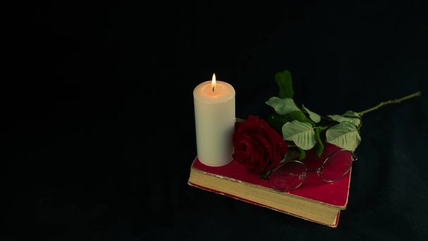 Una rosa roja con un libro y una vela encendida sobre un fondo negro. Desde arriba de la vela encendida con rojo fresco con una rosa en el libro sobre fondo negro. - Foto, imagen
