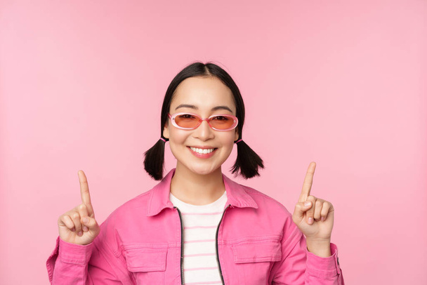 Nahaufnahme Porträt des modernen koreanischen weiblichen Modells, trägt Sonnenbrille, zeigt mit dem Finger nach oben, zeigt Werbung, Werbebanner, rosa Hintergrund - Foto, Bild