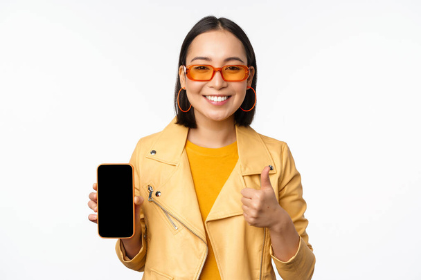 Belle fille coréenne, femme asiatique en lunettes de soleil, montrant l'interface de l'application smartphone, pouces levés, recommandant l'application de téléphone mobile, fond blanc - Photo, image